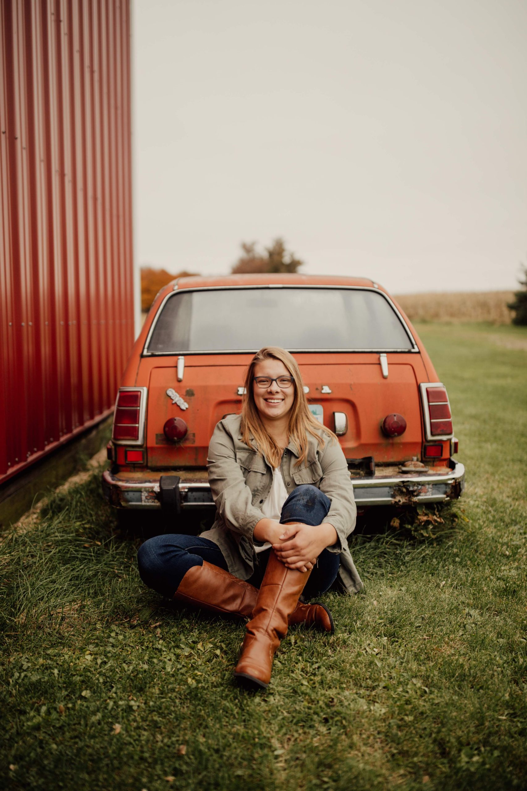 Outdoor portrait photographed in a rural setting with a vintage orange station wagon, red barn, and open farmland in the background.